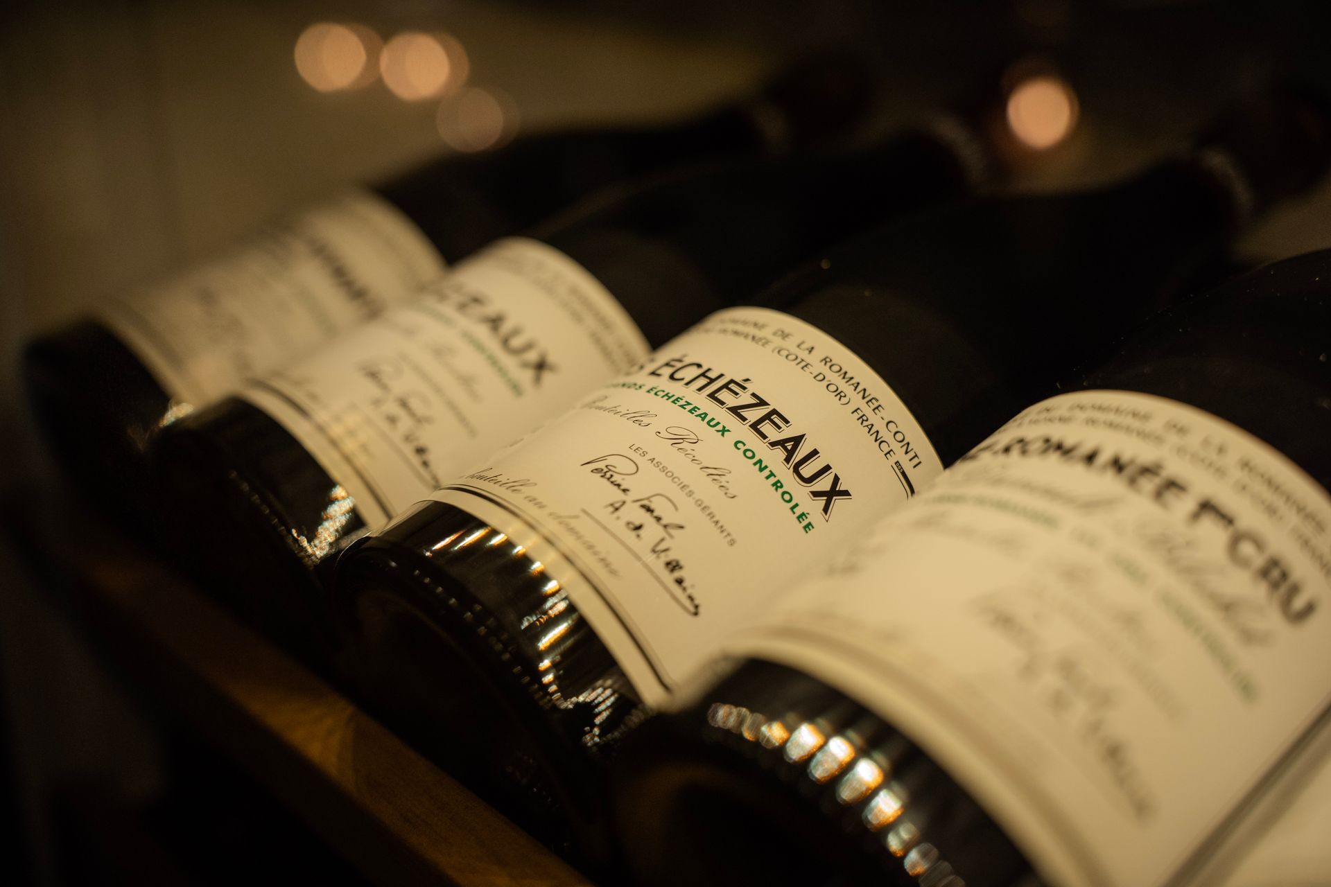 Een rij vintage wijnflessen met leesbare etiketten, waaronder Echezeaux, staat op een rek in een schemerige, sfeervolle kelder.
