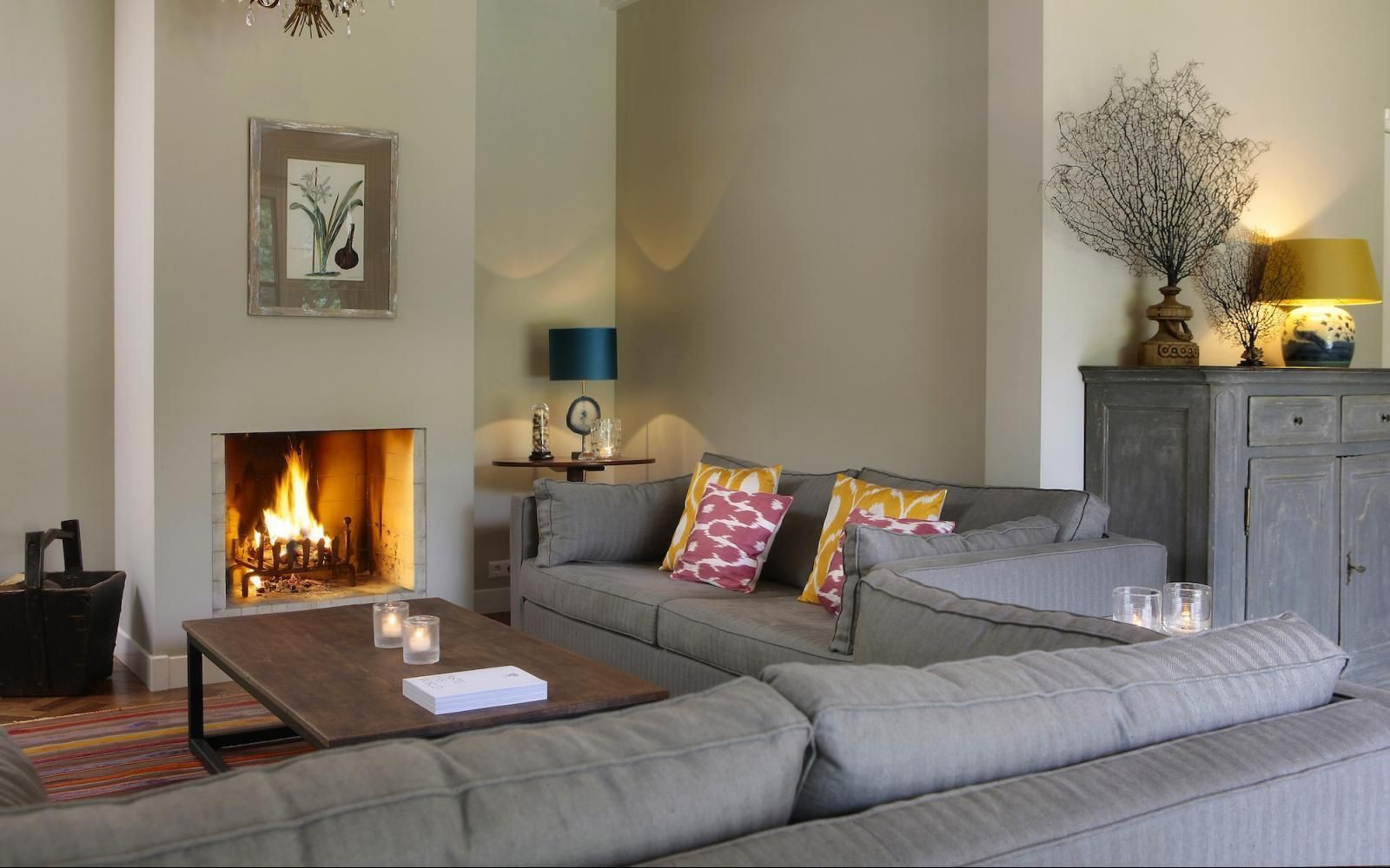 Een woonkamer met een brandende open haard, een grote grijze hoekbank, een houten salontafel en een kast met een lamp.