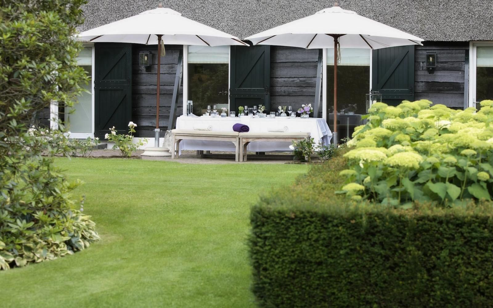 Een eettafel gedekt voor een maaltijd in de buitenlucht onder twee witte parasols op een gazon voor een huis met donkerhouten luiken.