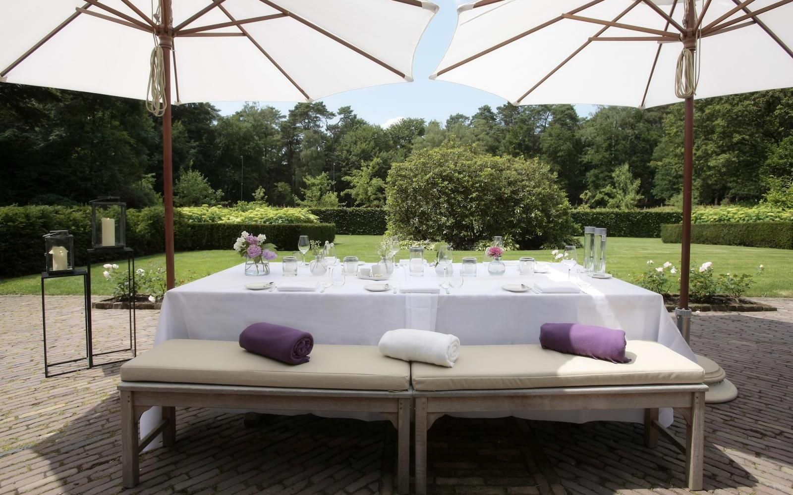 Auf einer mit Kopfsteinpflaster belegten Terrasse mit Blick auf einen grünen Garten steht unter zwei Sonnenschirmen ein weiß gedeckter Esstisch mit Bänken.