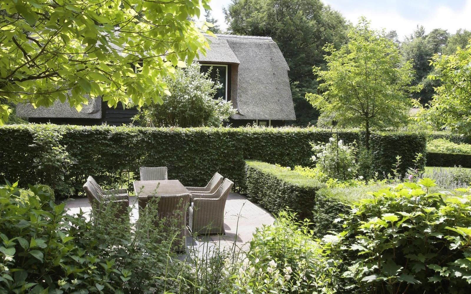 Een terras met rieten meubels in een weelderige, groene tuin met een hoge heg en een huis met een rieten dak op de achtergrond.