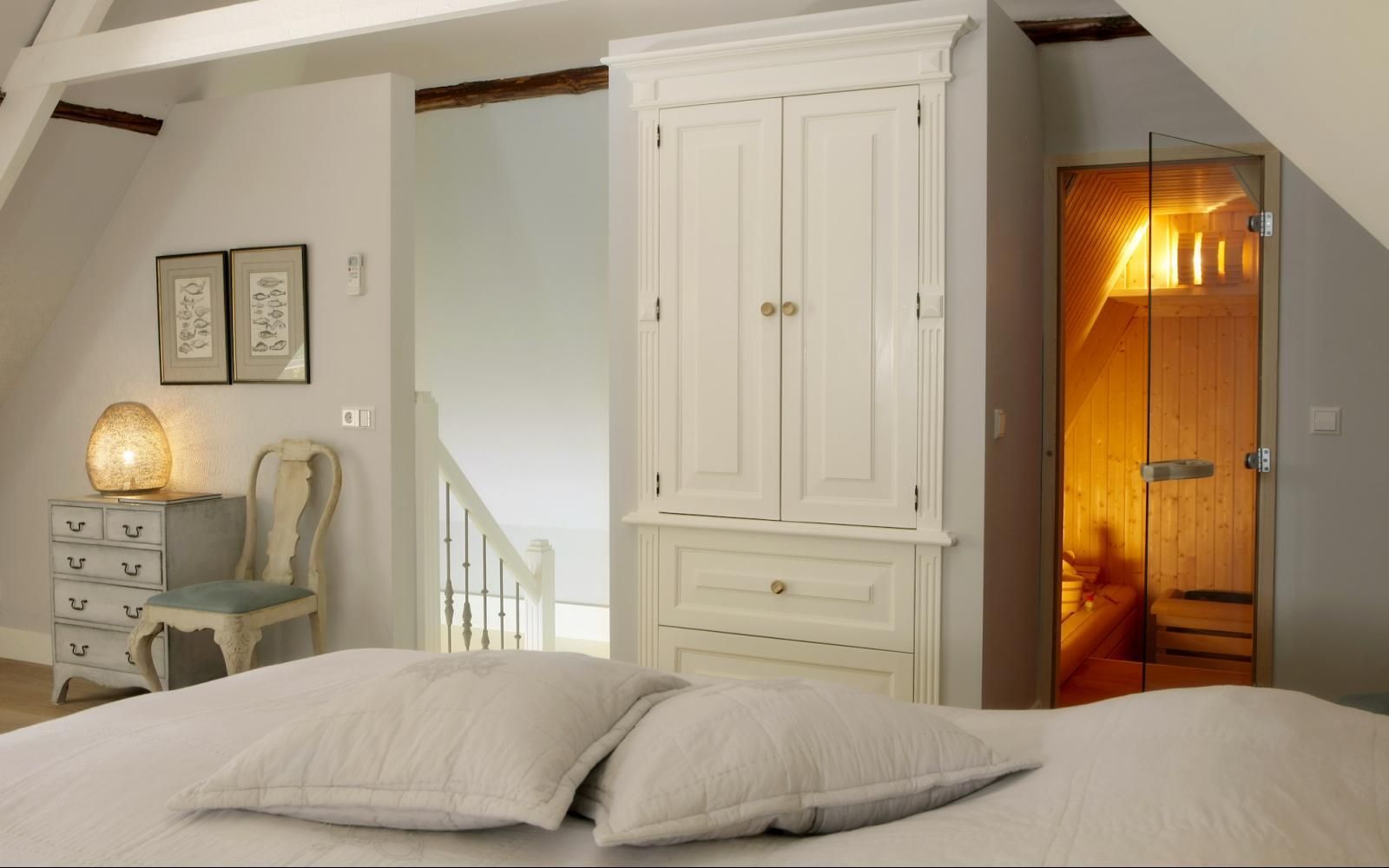 Ein Schlafzimmer mit einem weißen Kleiderschrank, einem kleinen Schreibtisch und Stuhl sowie einer Tür, hinter der sich eine helle, holzgetäfelte Sauna befindet.