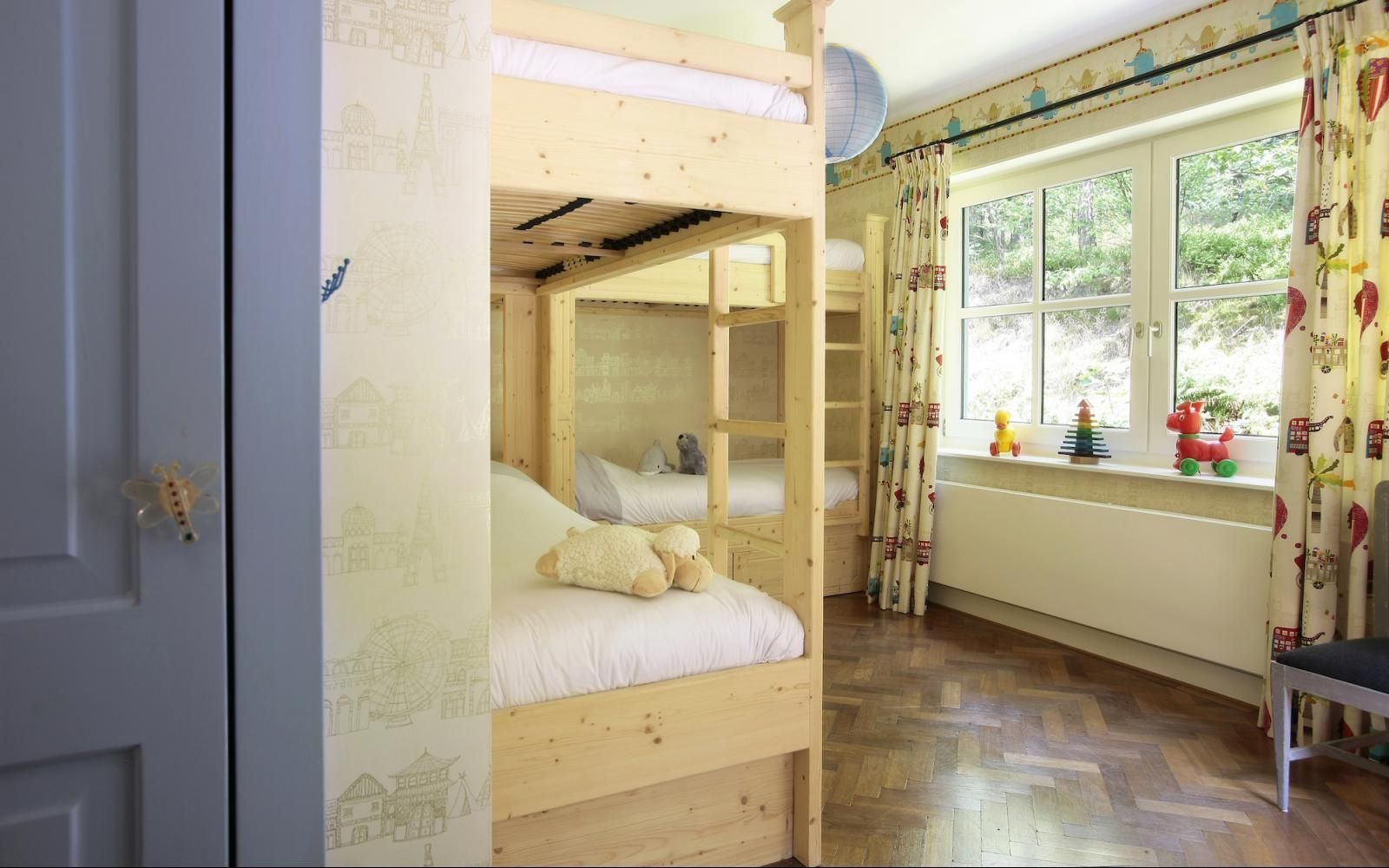 Een stapelbed van licht hout met wit beddengoed en een knuffeldier in een slaapkamer met een raam, gordijnen en een houten vloer.