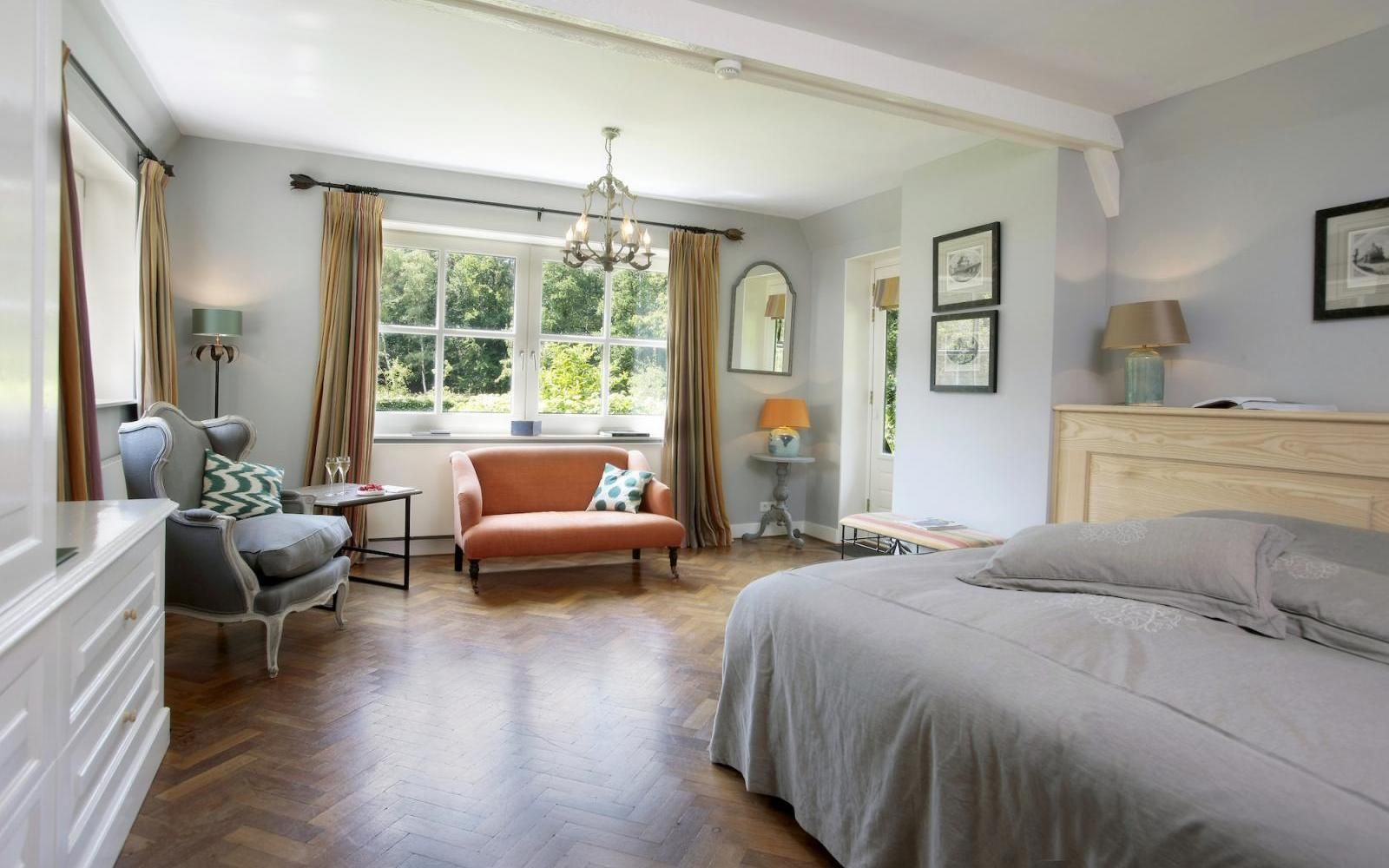 Een lichte, ruime slaapkamer met visgraatparket, een groot bed, een grijze fauteuil en een kleine oranje bank bij het raam.