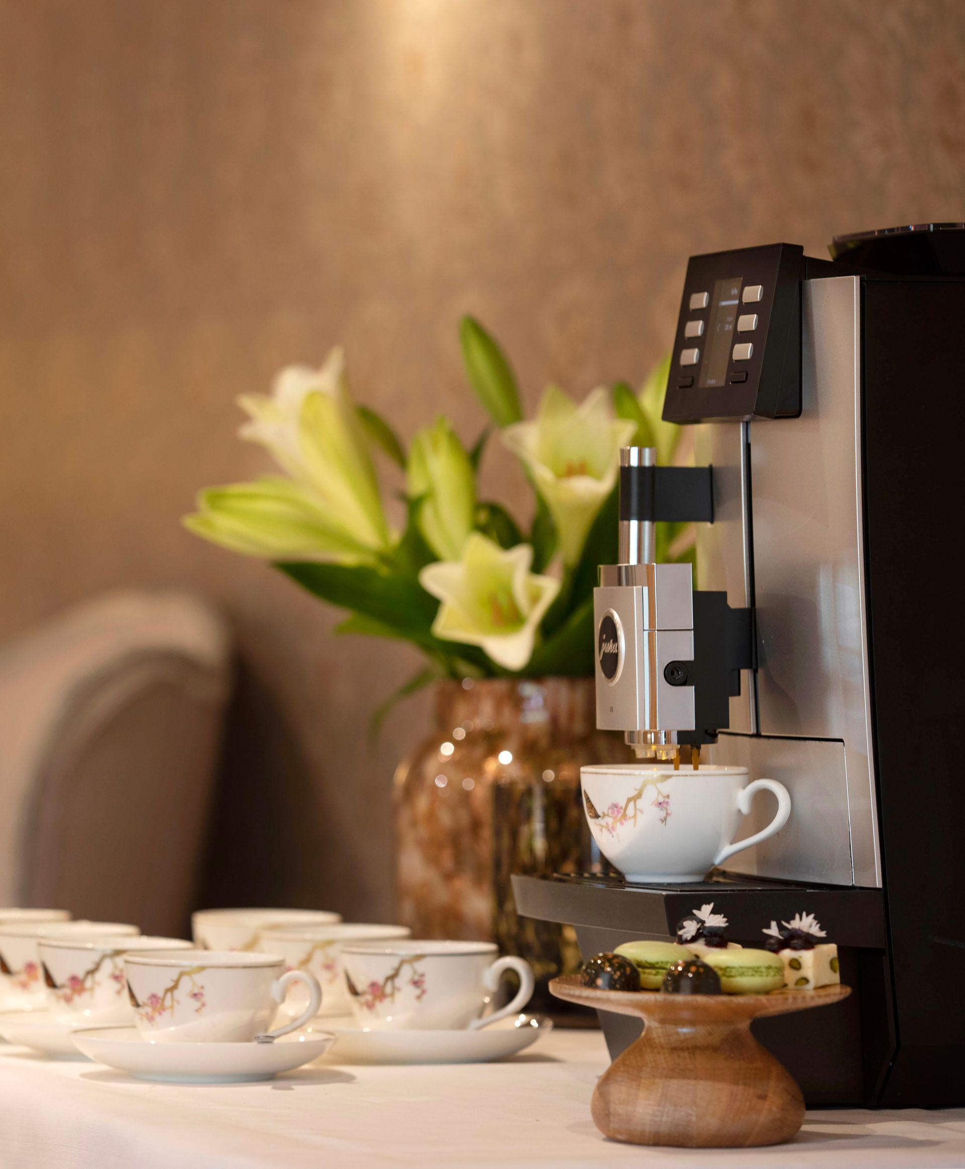 Een koffiemachine zet koffie in een kopje met een bloemenmotief, vlakbij witte lelies in een vaas, macarons en lege kopjes op een tafel.