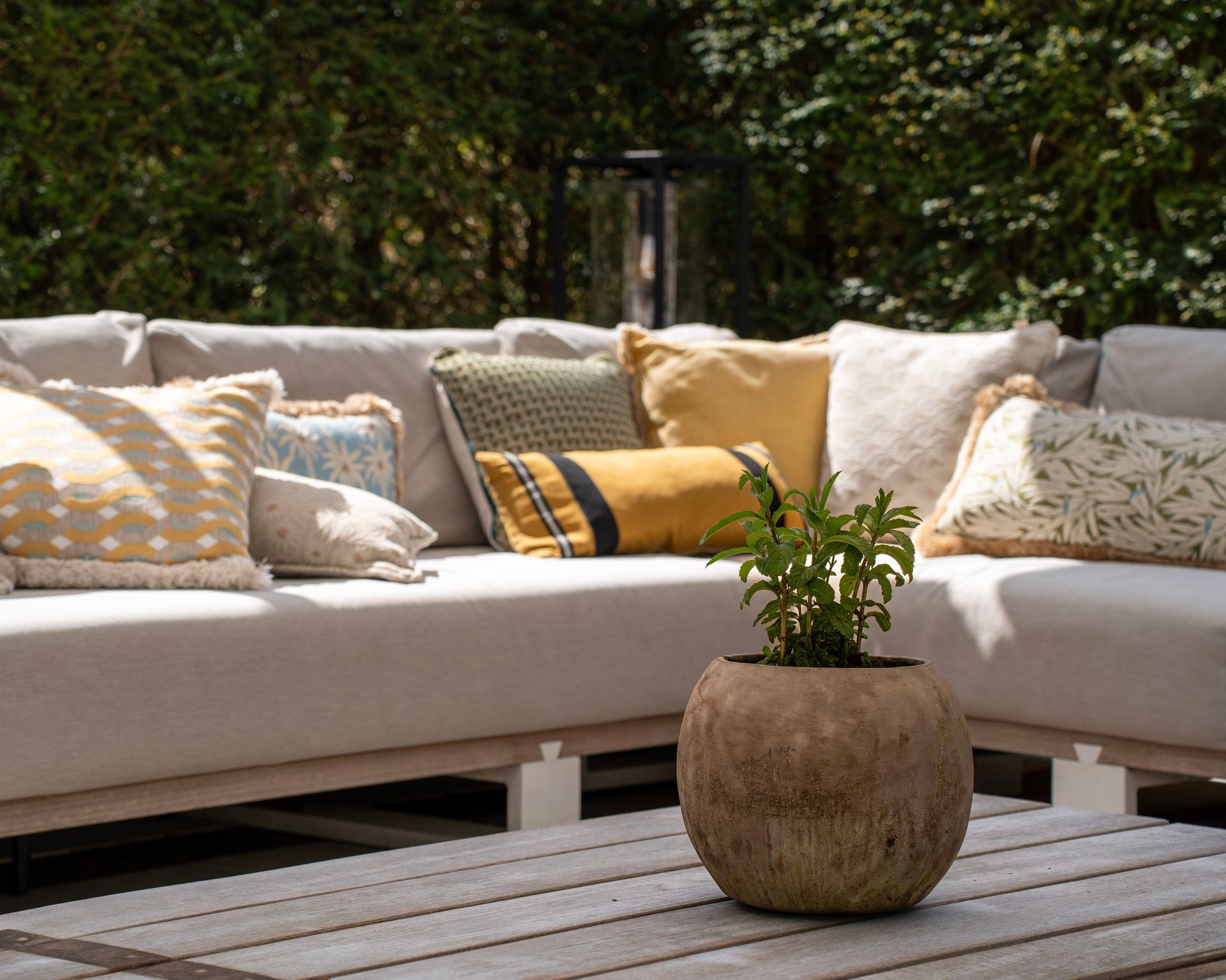 Een lichtgekleurde modulaire buitenbank met diverse sierkussens en een ronde plantenbak op een verweerde houten tafel.