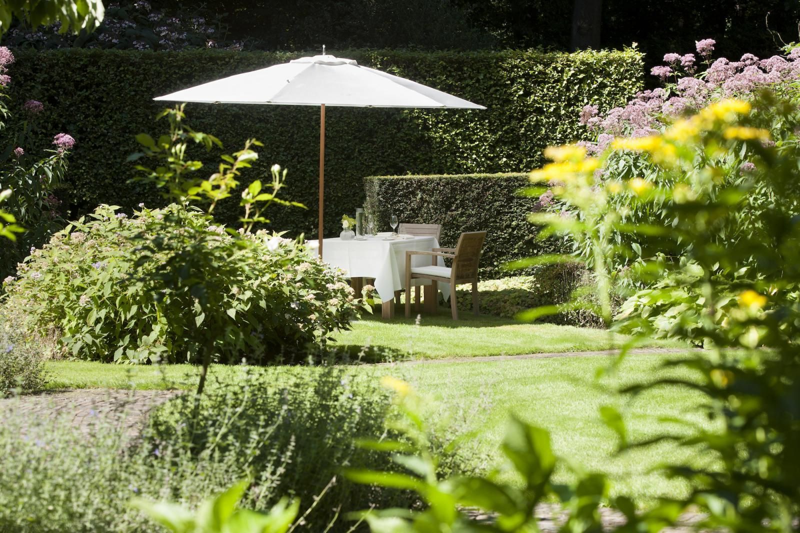 Een witte parasol biedt schaduw aan een tafel die gedekt is voor het diner in een weelderige, groene, zonovergoten tuin.