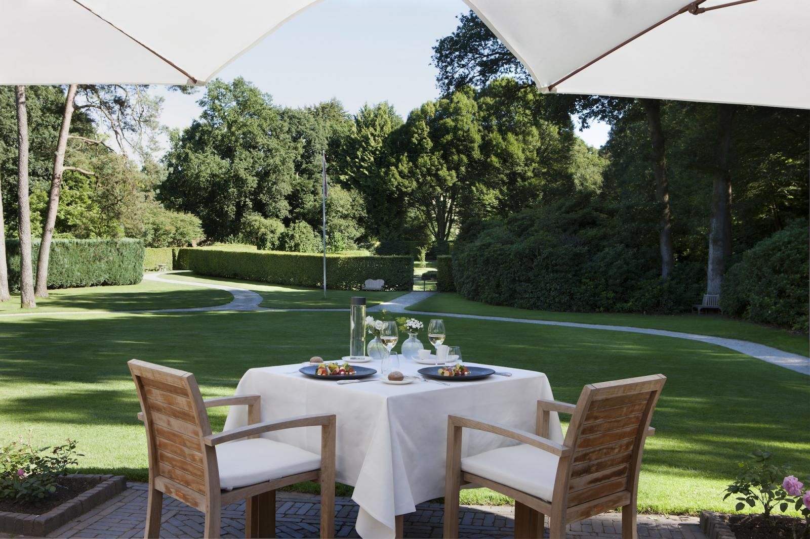 Op een terras met uitzicht op een weelderig groen gazon onder witte parasols staat een tafel gedekt voor twee personen met een wit tafelkleed.