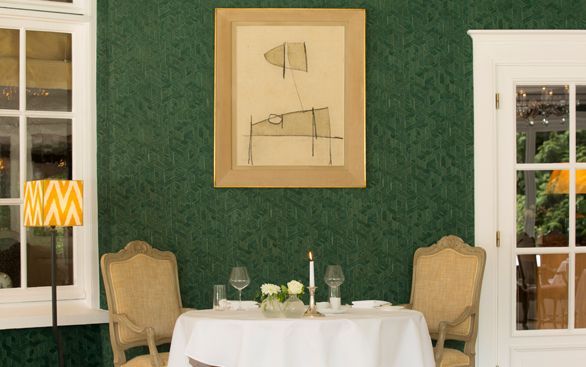 Een gedekte tafel voor twee met stoelen, een wit tafelkleed en minimalistische kunst op een donkergroene behangmuur.