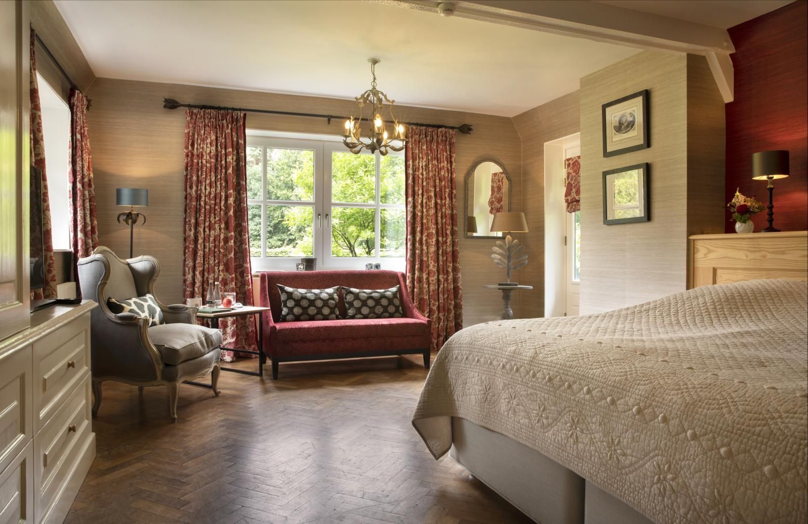 Ein geräumiges Schlafzimmer mit einem großen Bett, einem roten Zweisitzer-Sofa am Fenster mit gemusterten Vorhängen und einem grauen Sessel.