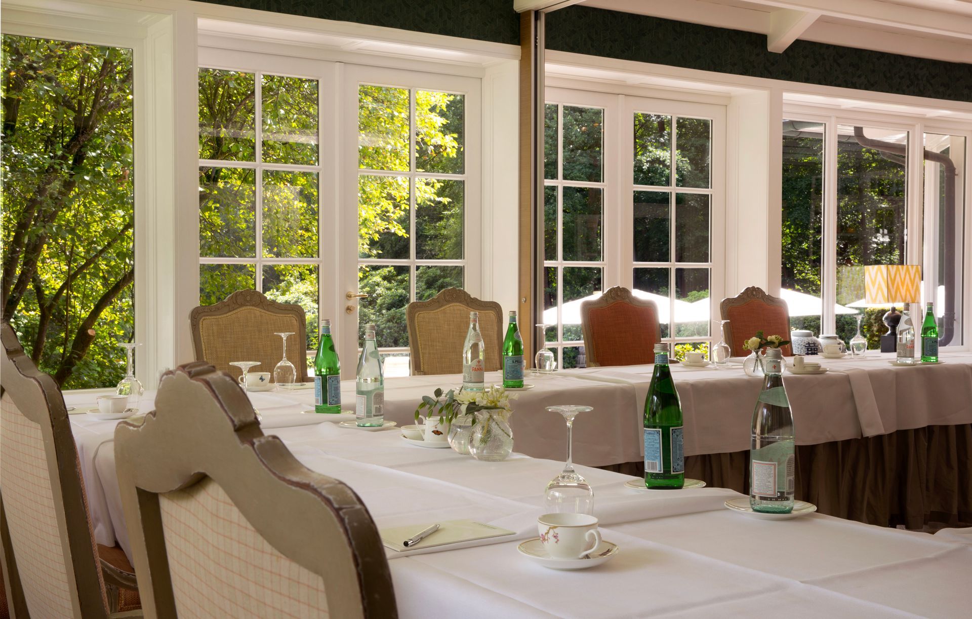 Paarse blauweregenbloemen hangen over een boog die een zonnig terras omlijst, met tafels, stoelen en witte parasols.