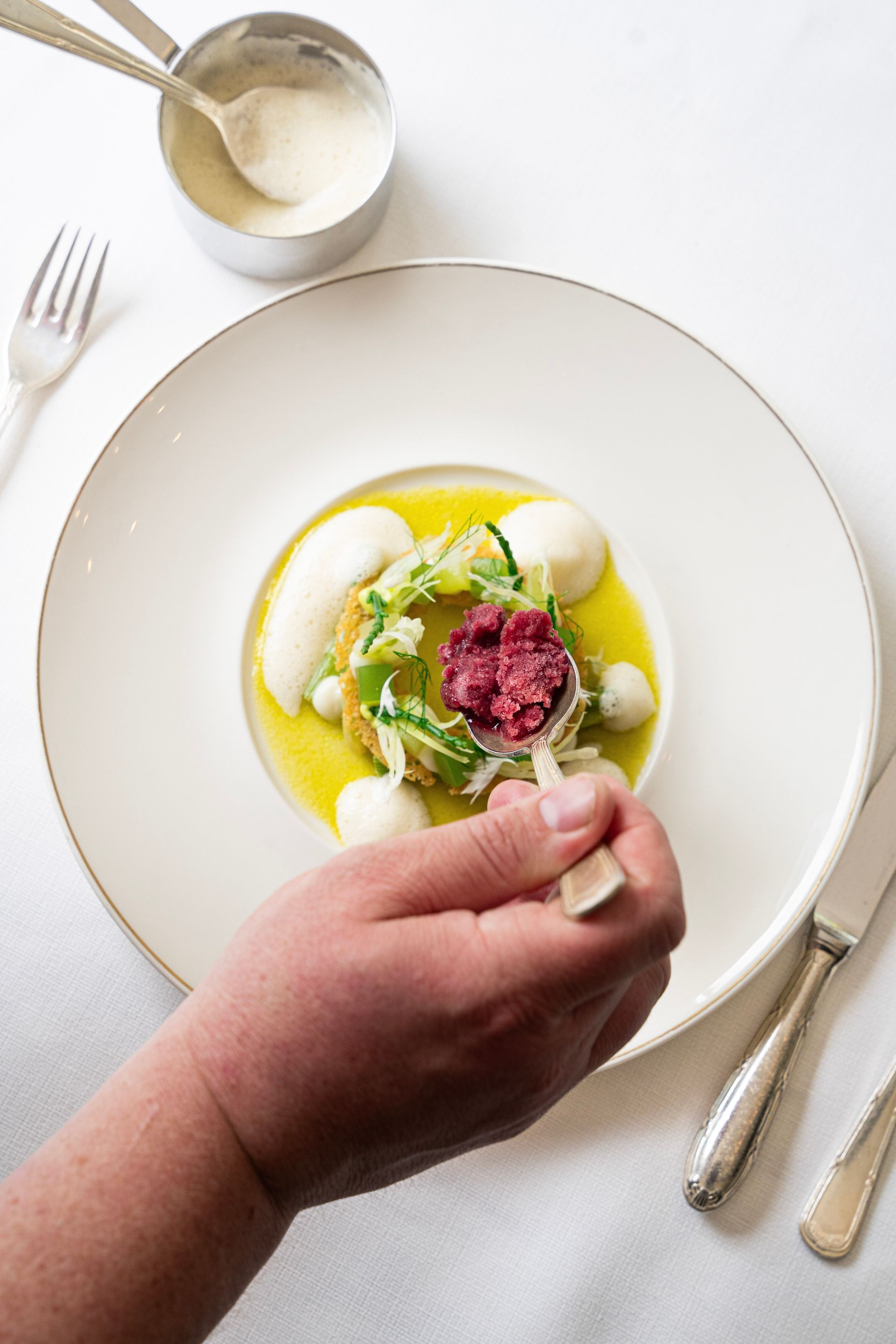Met een lepel schept iemand een kleurrijk, gastronomisch gerecht met groene bladgroenten, olie en een rode vulling in het midden op een wit gedekte tafel.