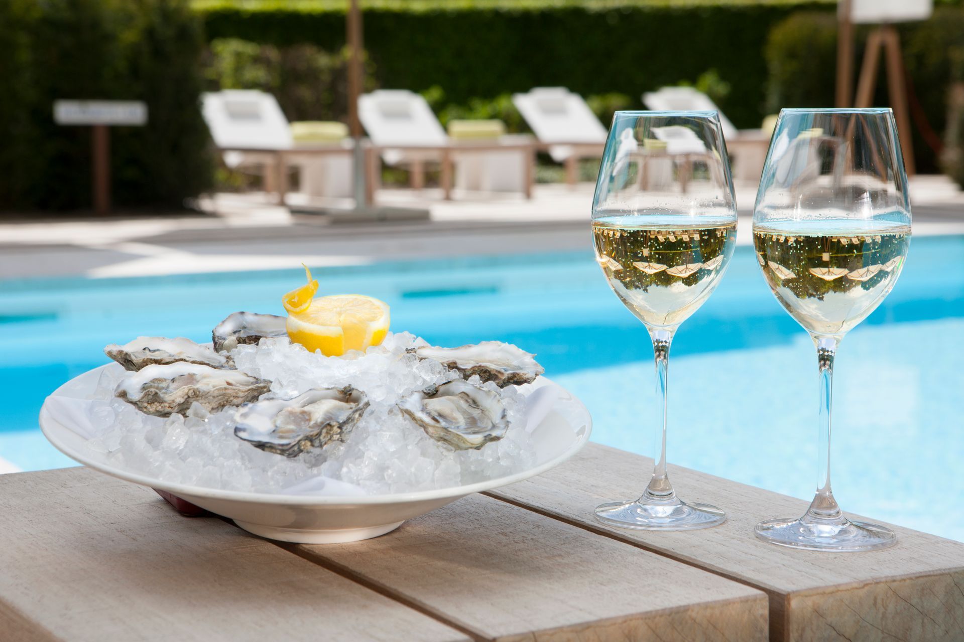 Een schaal met verse oesters op ijs met citroen, naast twee glazen witte wijn op een terras bij het zwembad.
