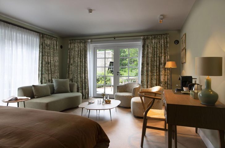 Een hotelkamer met een bed, een saliegroene bank, een salontafel, fauteuils en een houten bureau met een lamp bij glazen deuren.