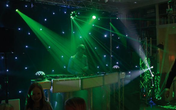 Een dj draait muziek op een feest met felgroene podiumverlichting, spotlights en een sterrenhemel als achtergrond.