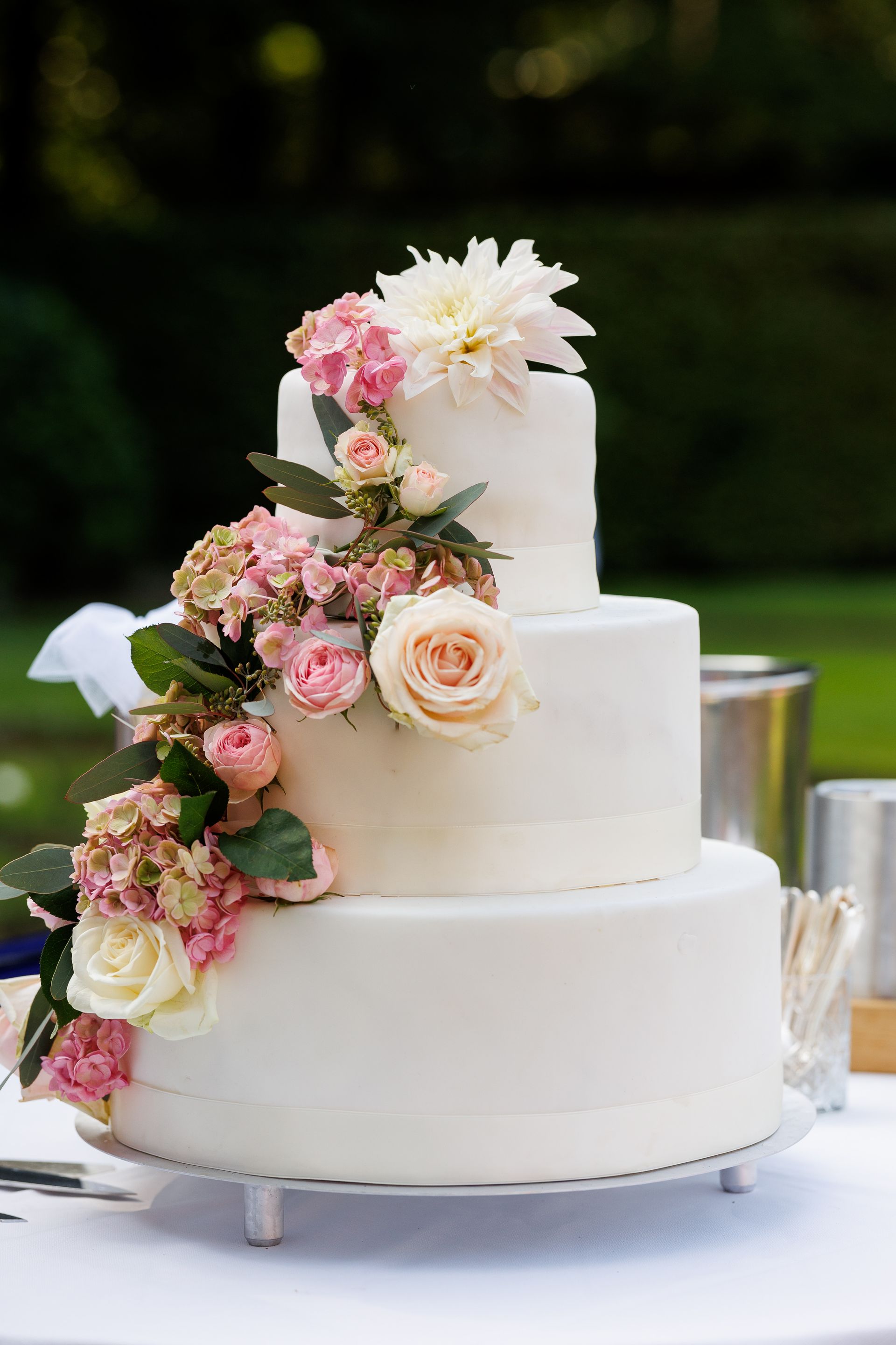 Een witte bruidstaart met drie lagen, versierd met een waterval van roze en witte rozen en hortensia's.