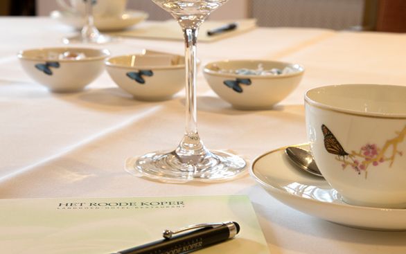 Een formele tafelsetting met theekopjes met vlindermotieven, kommen, een wijnglas, een pen en een notitieblok.