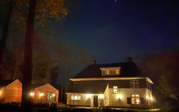 Een huis met verlichte ramen en buitenverlichting 's nachts, omgeven door bomen onder een donkere hemel.