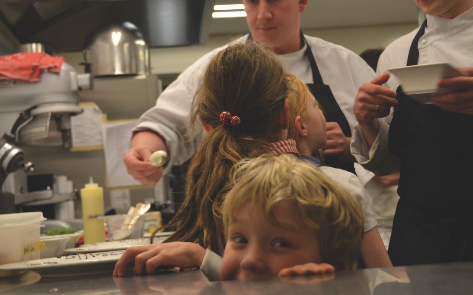 Mensen in koksuniformen praten met kinderen die zich rond een aanrecht hebben verzameld, waarbij een van de kinderen over de rand gluurt.