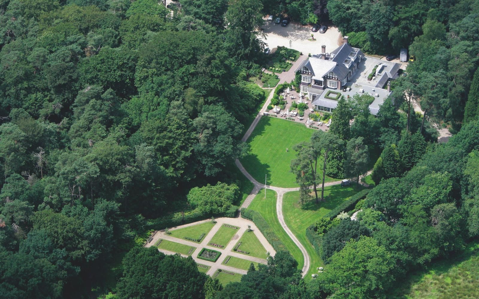 Luchtfoto van een huis verscholen in een dicht bos, met een rond gazon en een geometrisch tuinpad.