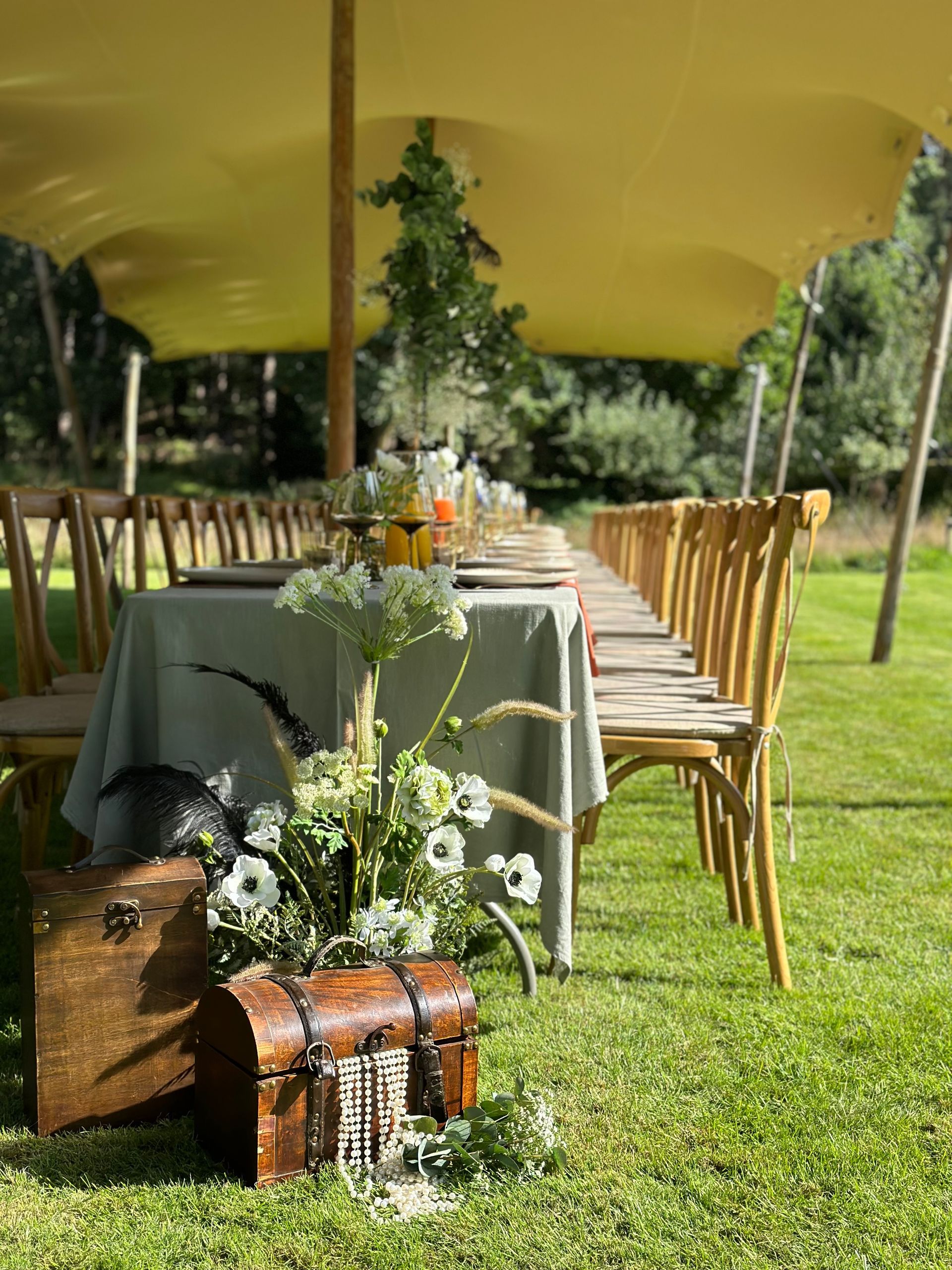 Een lange eettafel, gedekt voor een buitenevenement onder een beige luifel, met een rustieke houten kist en een bloemstuk in het midden.