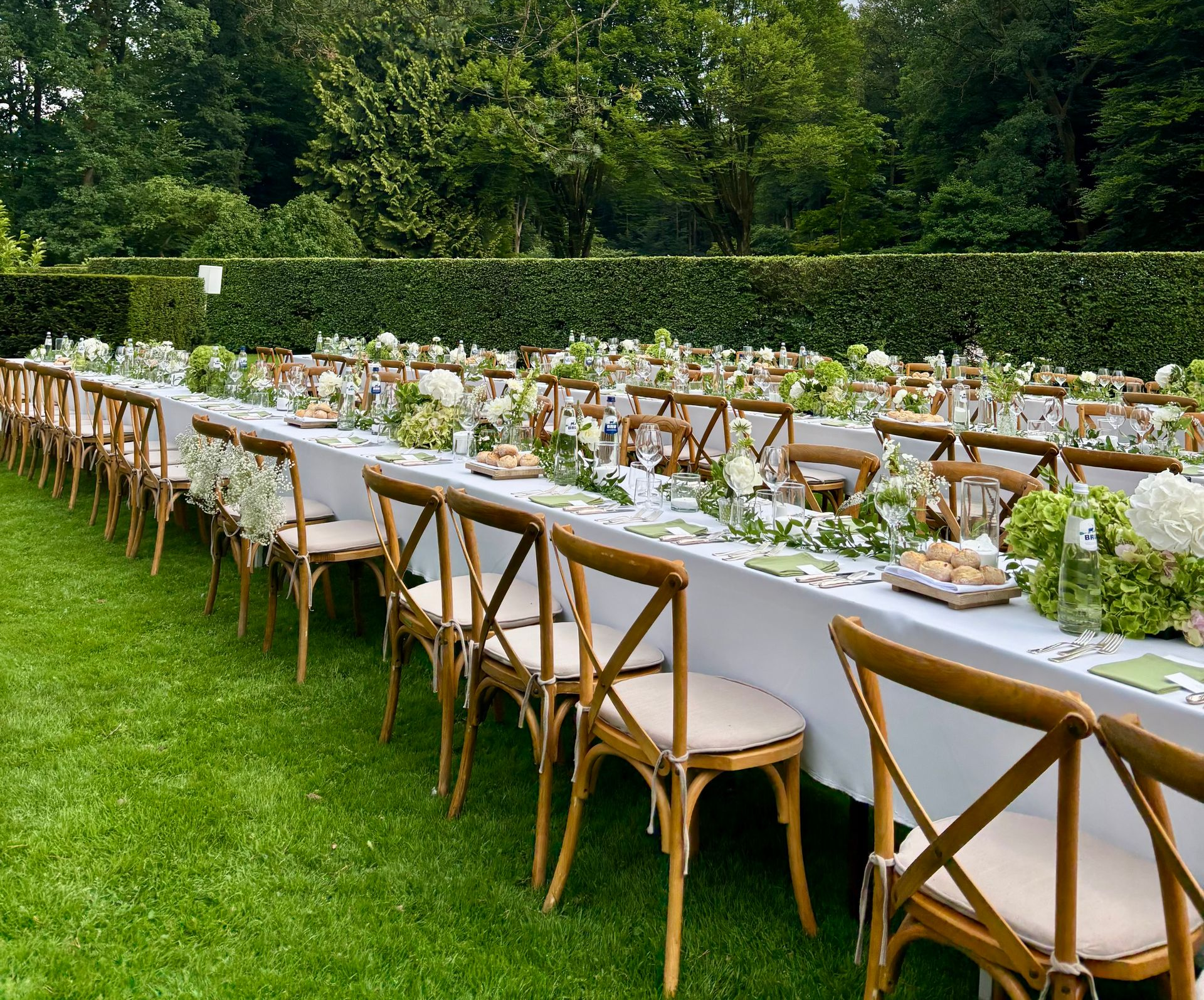 Een lange feesttafel, gedekt voor een evenement in de buitenlucht op een grasveld, met houten stoelen met kruisvormige rugleuningen en witte tafelkleden.