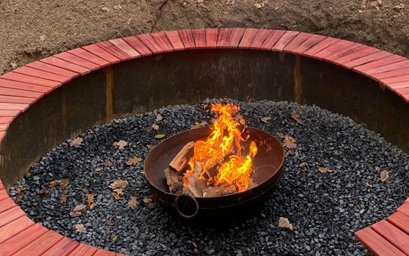 In een metalen schaal in een grindbak brandt een vuur, omringd door een ronde houten bank.