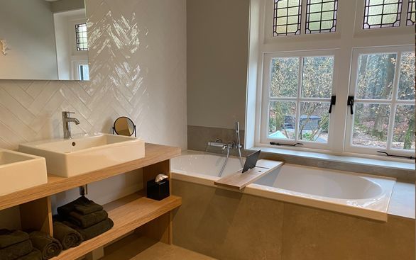 Moderne badkamer met een houten badmeubel, twee witte opbouwspoelbakken en een ingebouwd bad onder een raam met roedenverdeling.