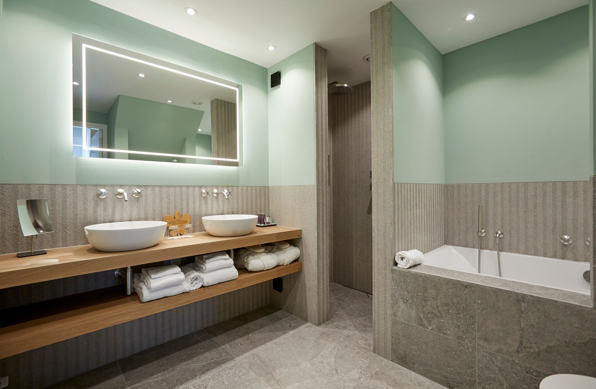 Moderne badkamer met dubbele wastafel, betegeld bad met natuurstenen tegels, zacht mintgroene muren en warme houten accenten.