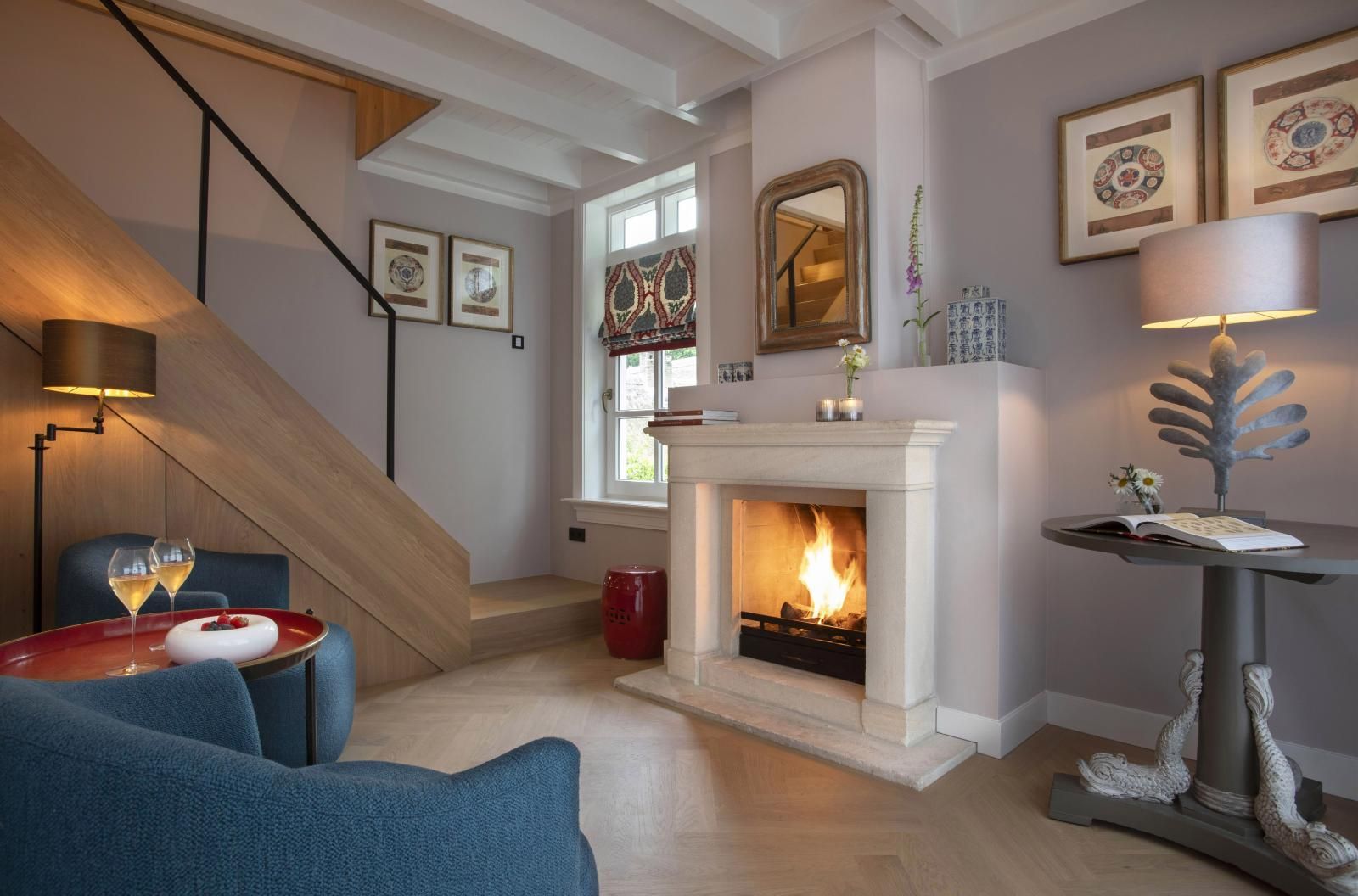 Een knusse kamer met een open haard, blauwe fauteuils, een houten trap en een tafellamp op een sokkel.