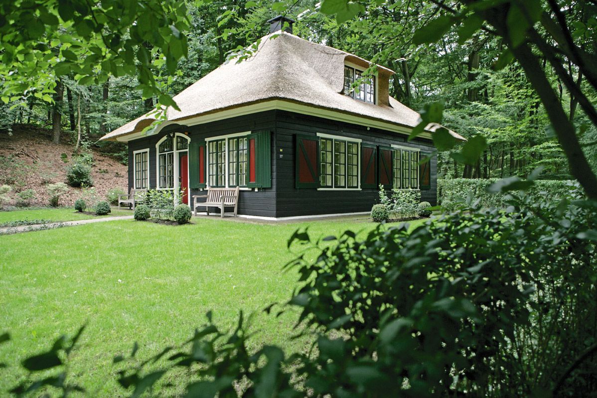 In een weelderige, groene open plek in het bos staat een donkerhouten huis met een rieten dak.