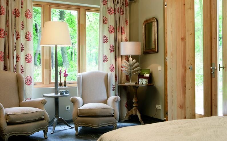 In einem gemütlichen Zimmer stehen zwei beige Sessel an großen Fenstern mit gemusterten Vorhängen, neben Steh- und Tischlampen.