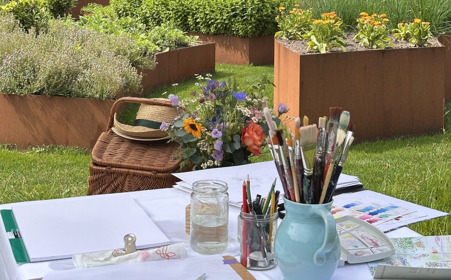 Teken- en schildersbenodigdheden, penselen in een blauwe kan, een waterkruik en een strohoed staan ​​uitgestald op een tafel in een zonnige, bloemrijke tuin.
