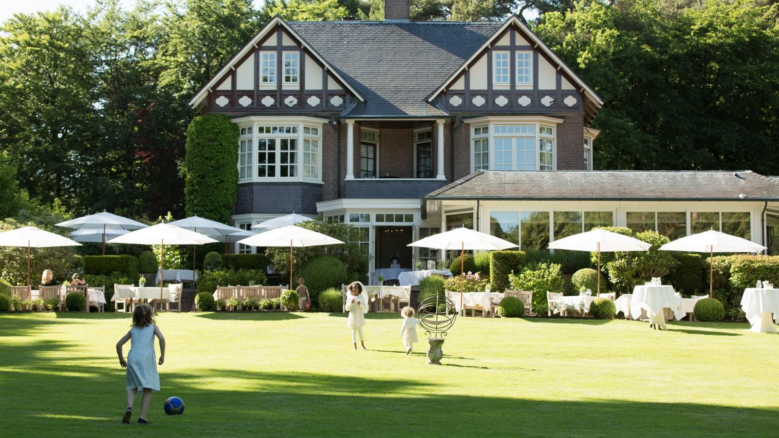 Een kind speelt met een bal op een zonnig gazon voor een groot huis met een terras dat is ingericht om buiten te eten.