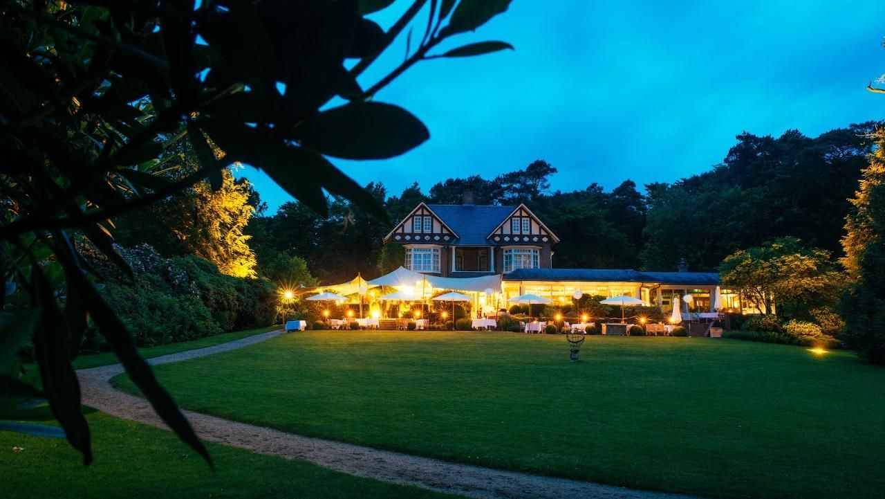 Een verlicht landhuis in de schemering, met een uitgestrekt groen gazon op de voorgrond en donkere bomen op de achtergrond.