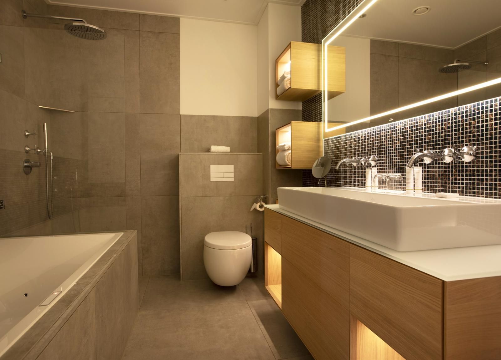 Een moderne badkamer met een wit bad, een wandtoilet, een houten badmeubel met een grote wastafel en een verlichte spiegel.