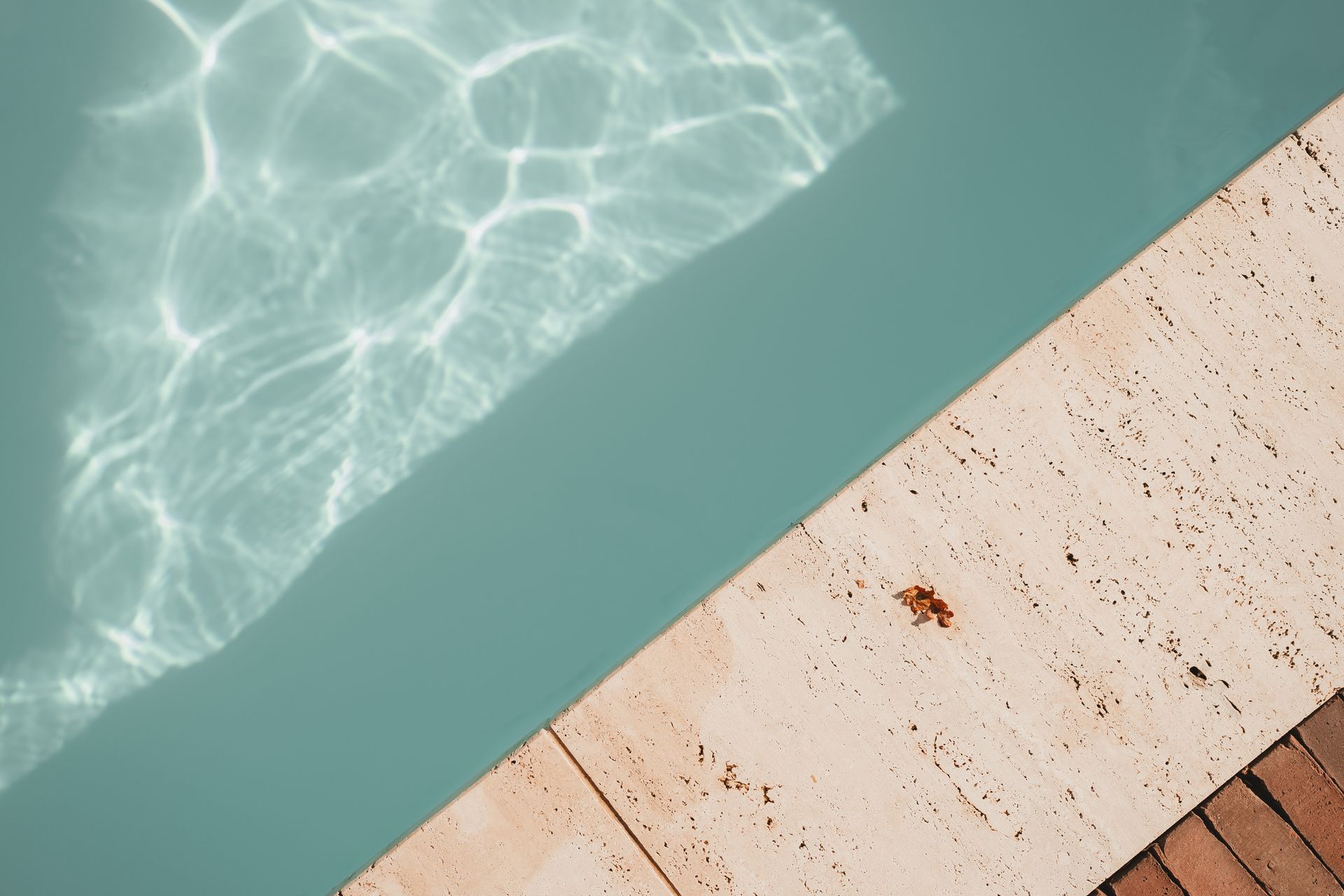 Een bovenaanzicht van turquoise zwembadwater met rimpelingen in het zonlicht, naast een terras van beige natuursteen met een bijzondere structuur.