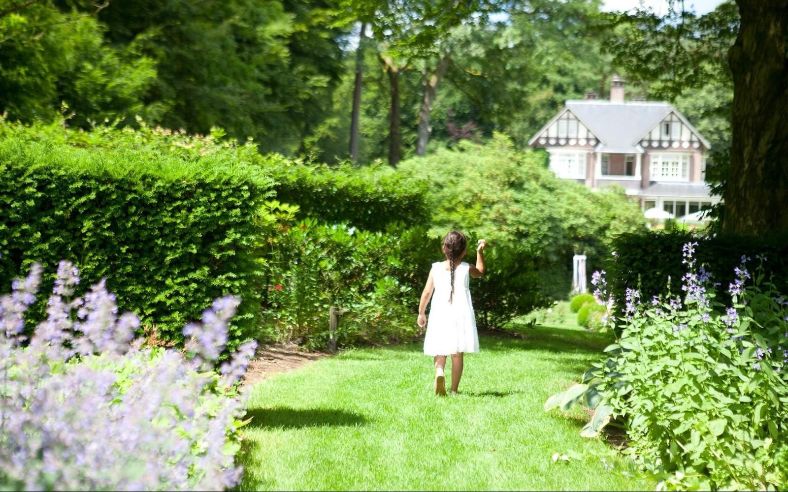Een persoon in een witte jurk loopt over een graspad naar een groot huis in een weelderige, groene tuin.