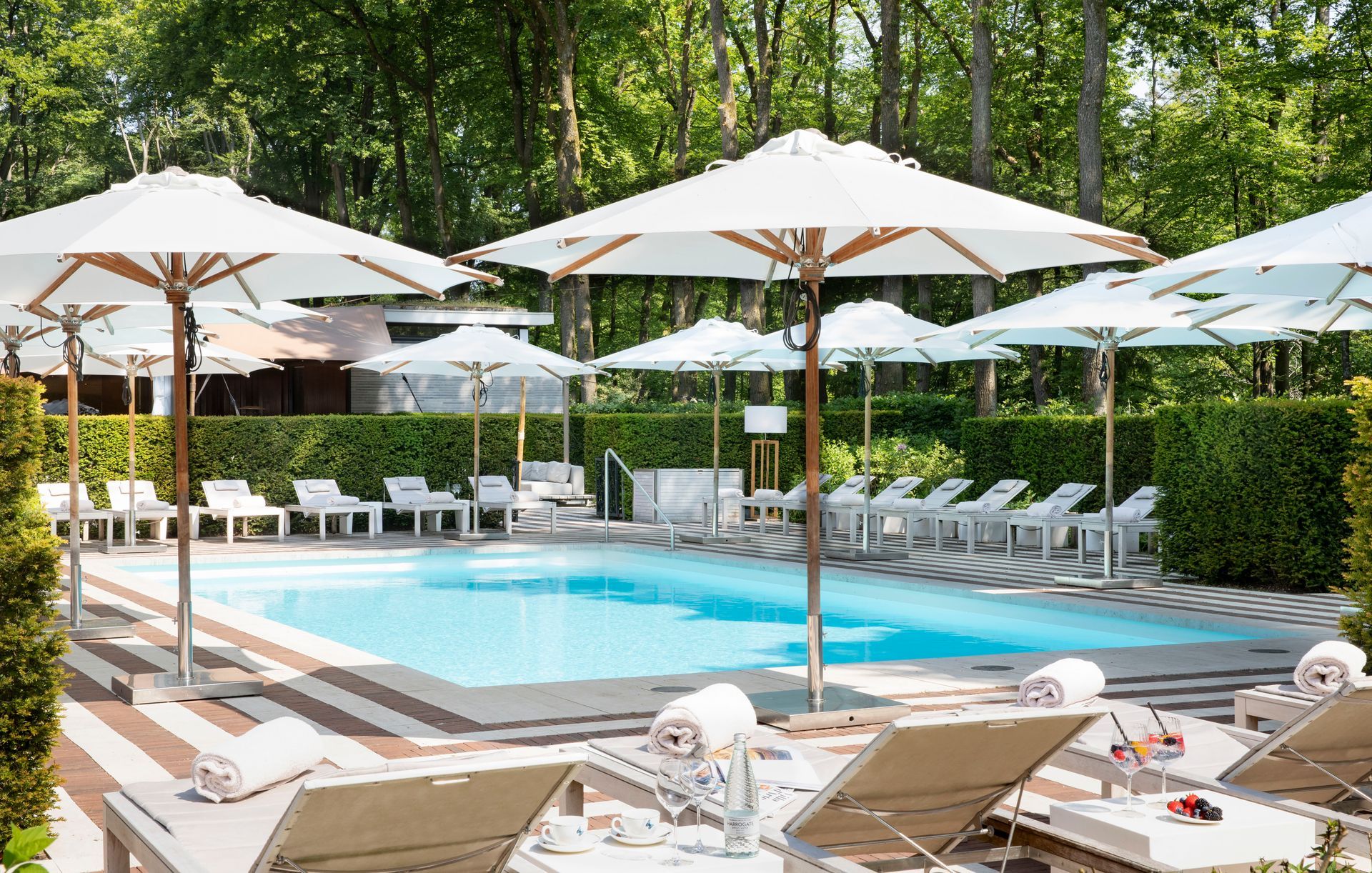 Een sereen zwembad, omgeven door witte ligstoelen en parasols, met op de achtergrond weelderige groene bomen.