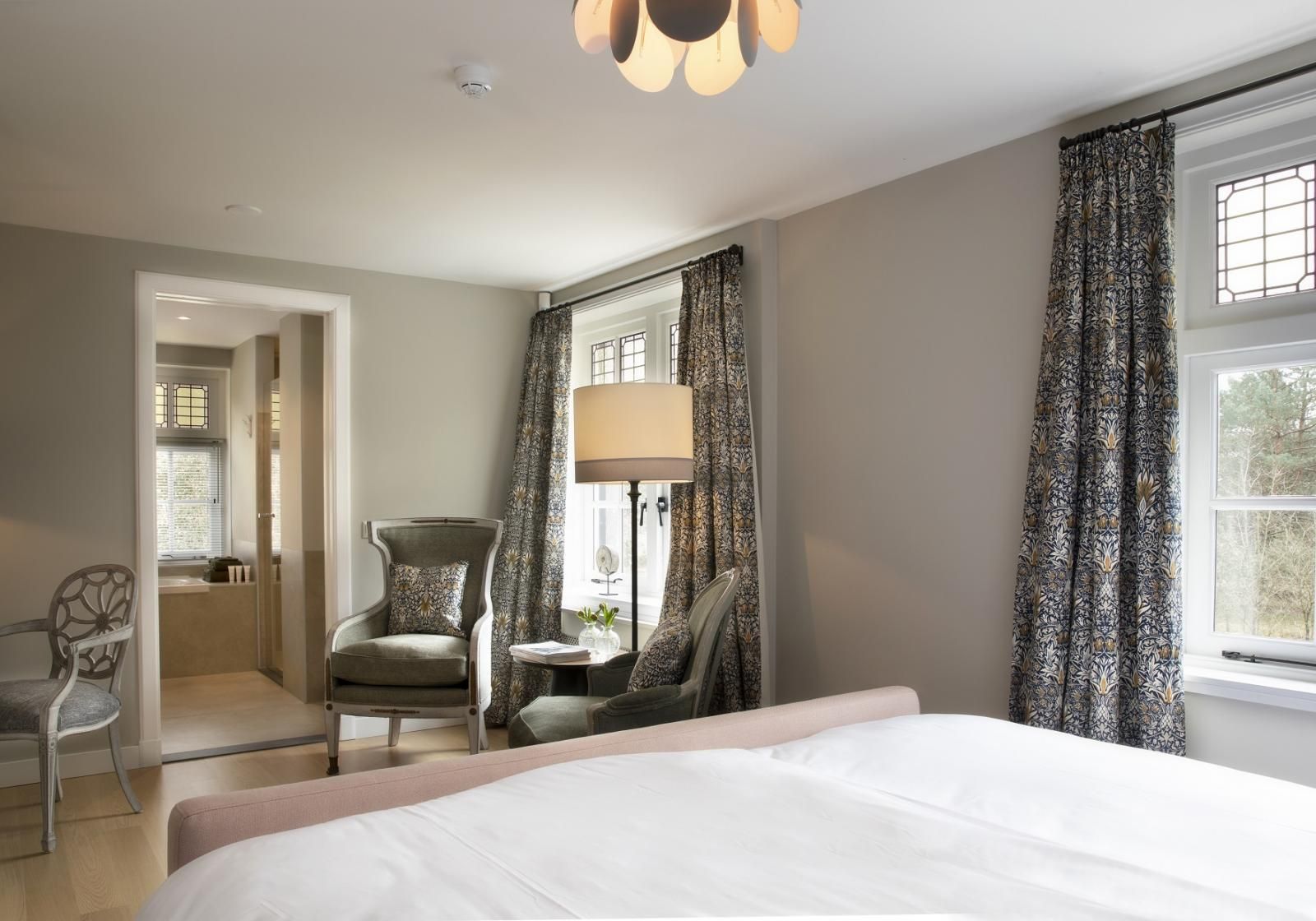Een neutraal ingerichte hotelkamer met een bed, een raam met gordijnen met een patroon, twee stoelen en een aangrenzende badkamer.