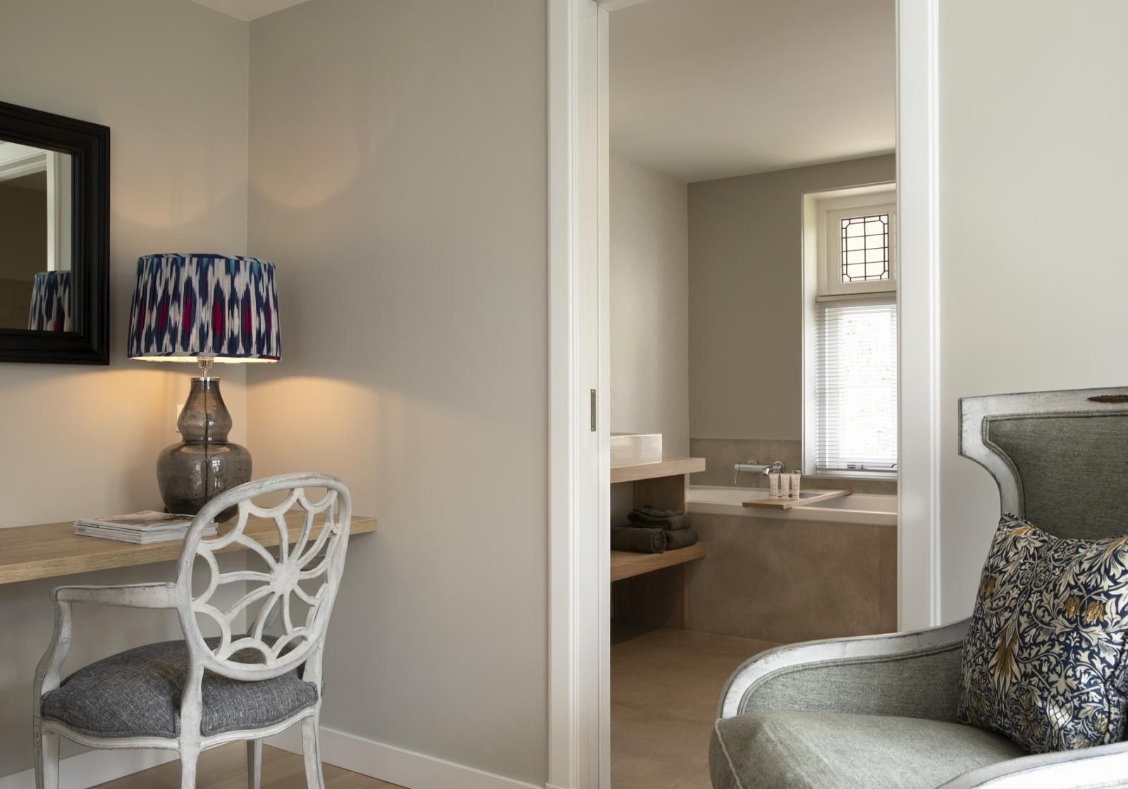 Een blik door een deuropening naar een badkamer vanuit een kamer met een bureau met een lamp en een fauteuil met een patroon.