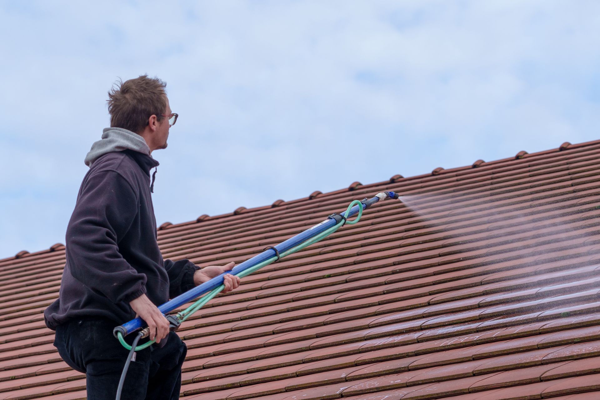 Une personne utilise un outil à long manche pour nettoyer un toit de tuiles sous un ciel nuageux.