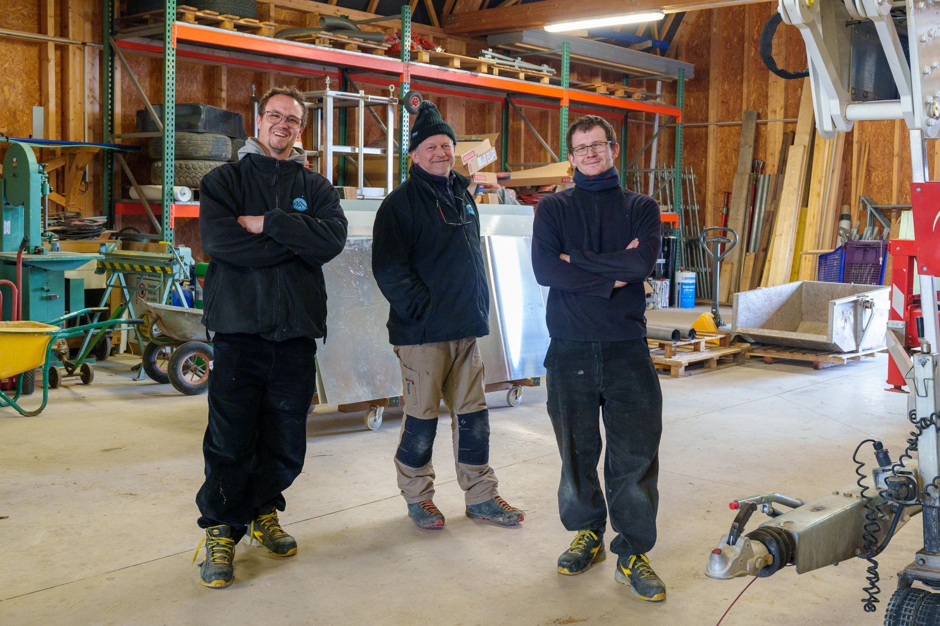 Trois personnes debout dans un atelier, les bras croisés, avec des outils et du matériel en arrière-plan.
