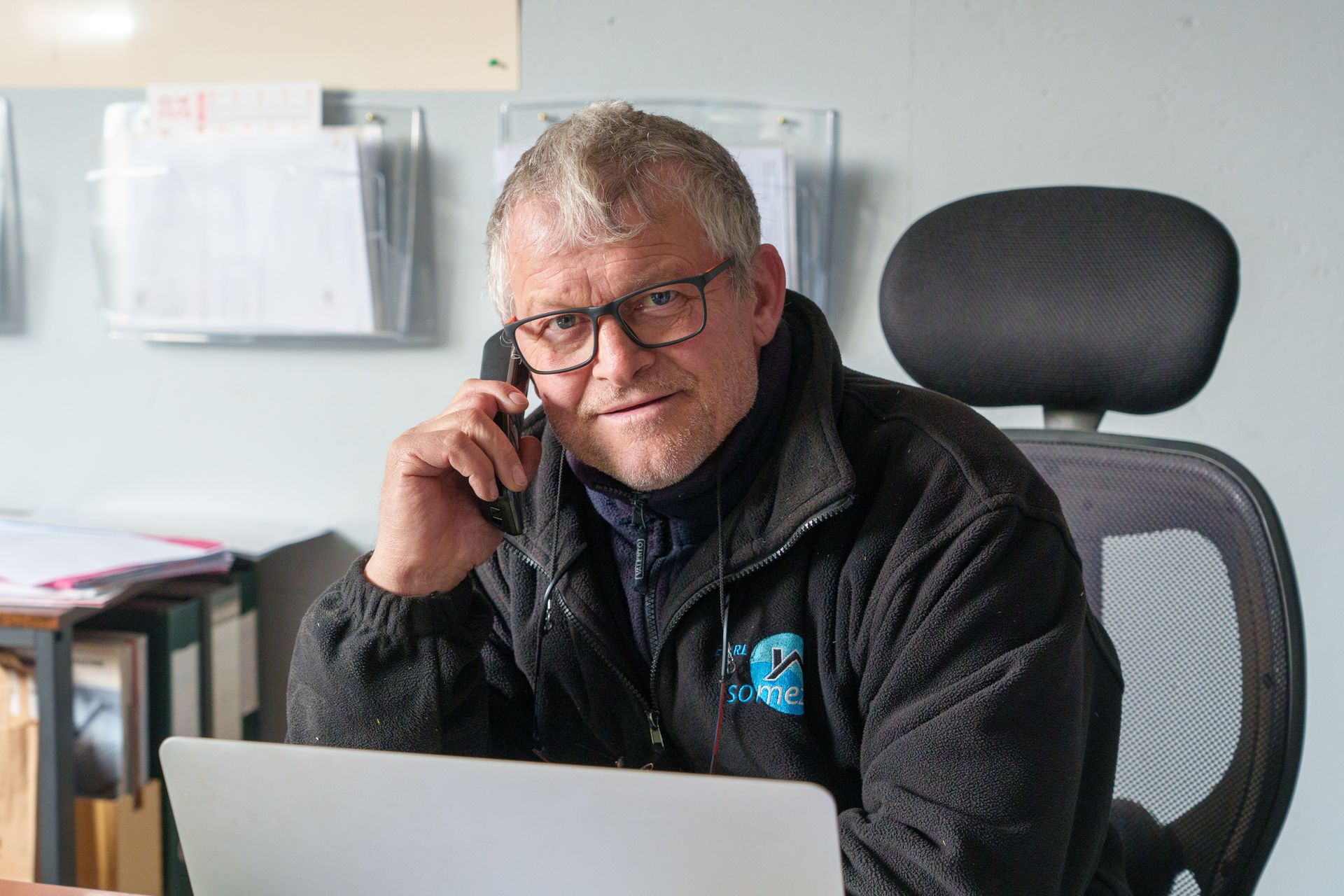 Un homme assis à un bureau, un téléphone à l'oreille, à côté d'un ordinateur portable, sur une chaise de bureau.