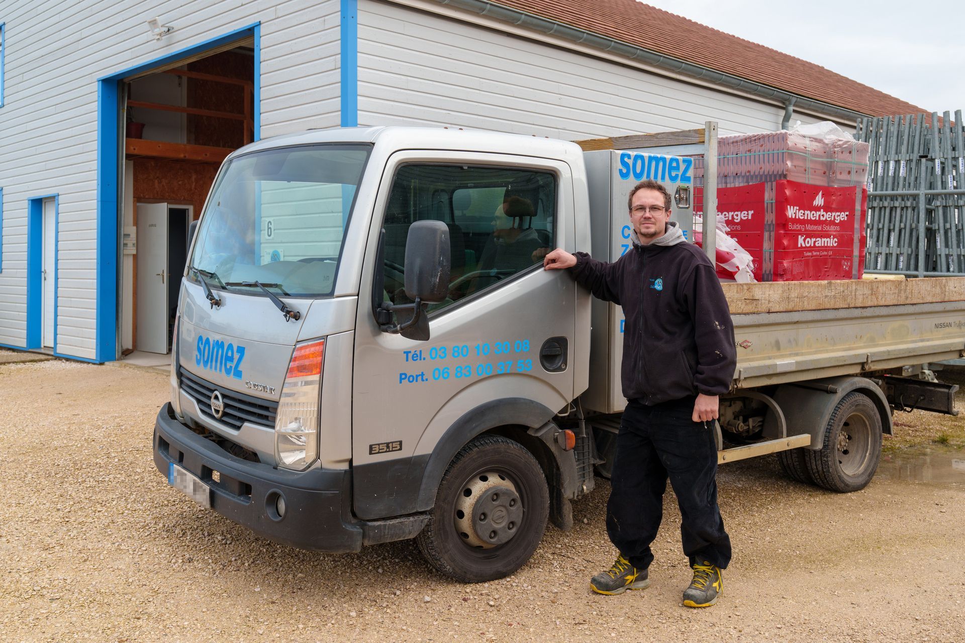 Un homme se tient à côté d'un camion plateau gris devant un garage-atelier.