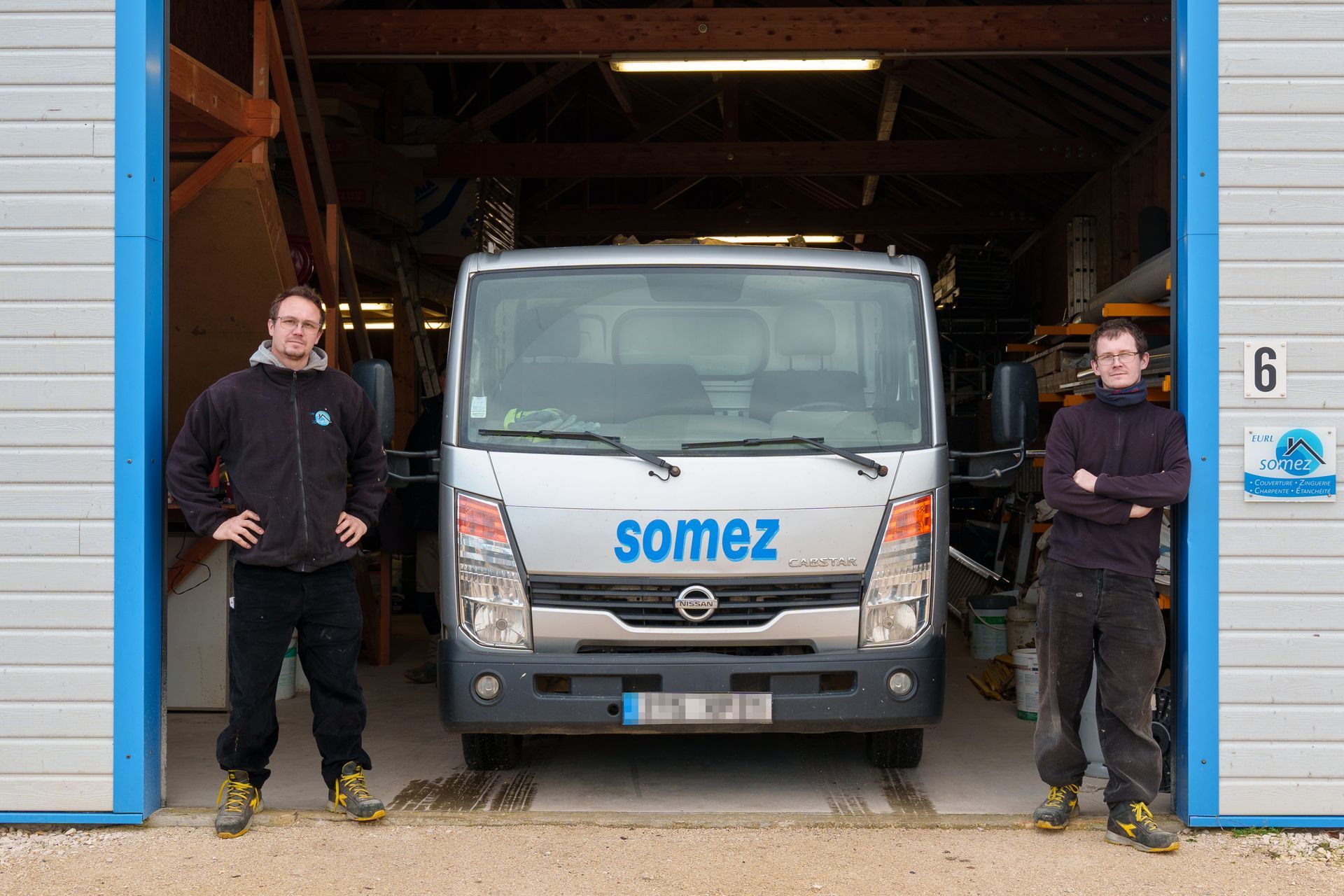 Deux ouvriers se tiennent debout à côté d'un camion Somez argenté dans l'embrasure d'une porte de garage.