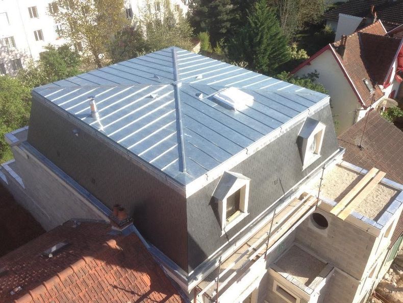 Vue aérienne d'une maison avec un toit en métal bleu et des lucarnes, située à proximité d'un chantier.