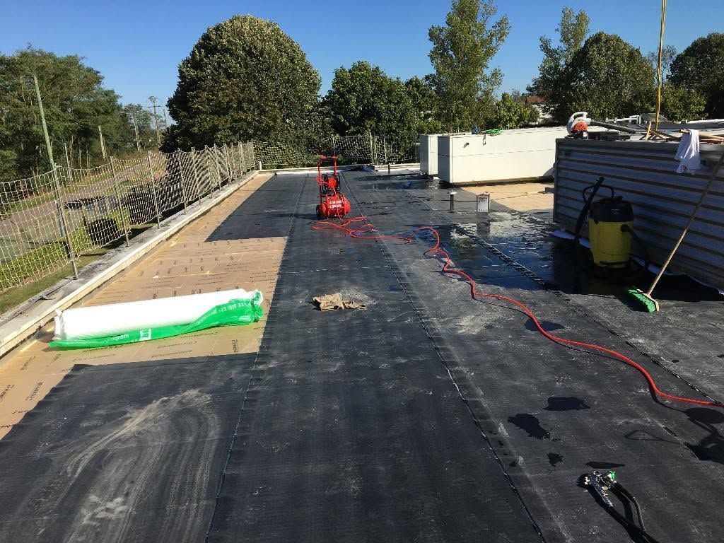 Toiture en réparation avec membrane noire, outils, rallonge électrique rouge et arbres en arrière-plan.