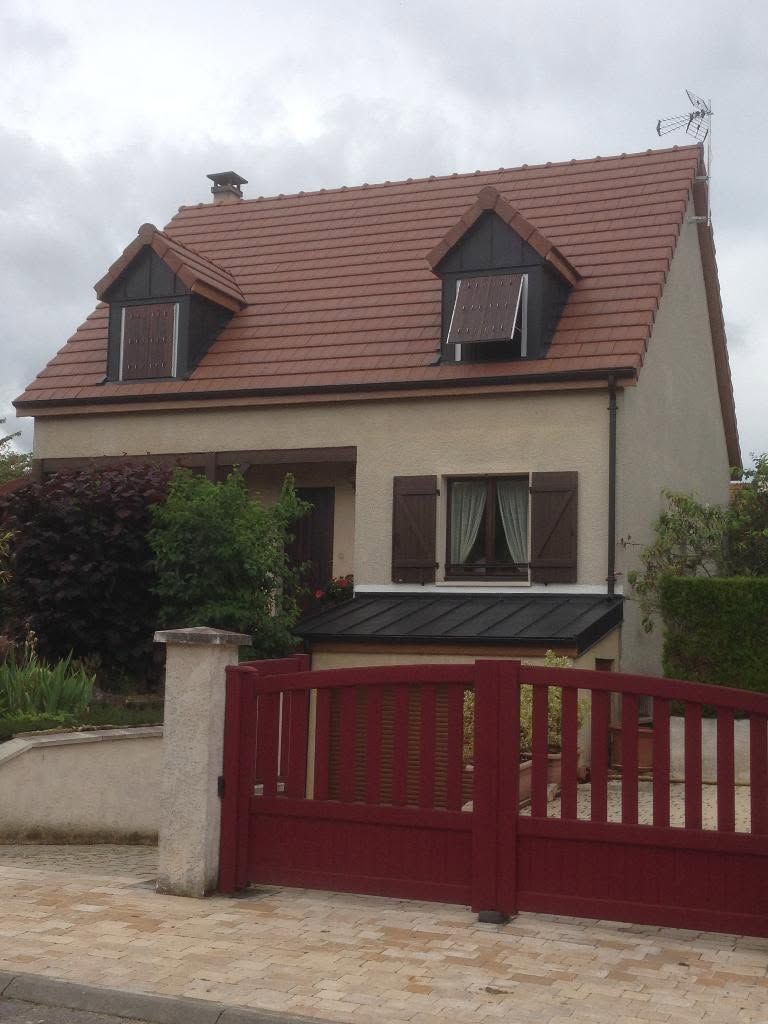 Maison à deux étages avec toit rouge, lucarnes et portail rouge en façade.