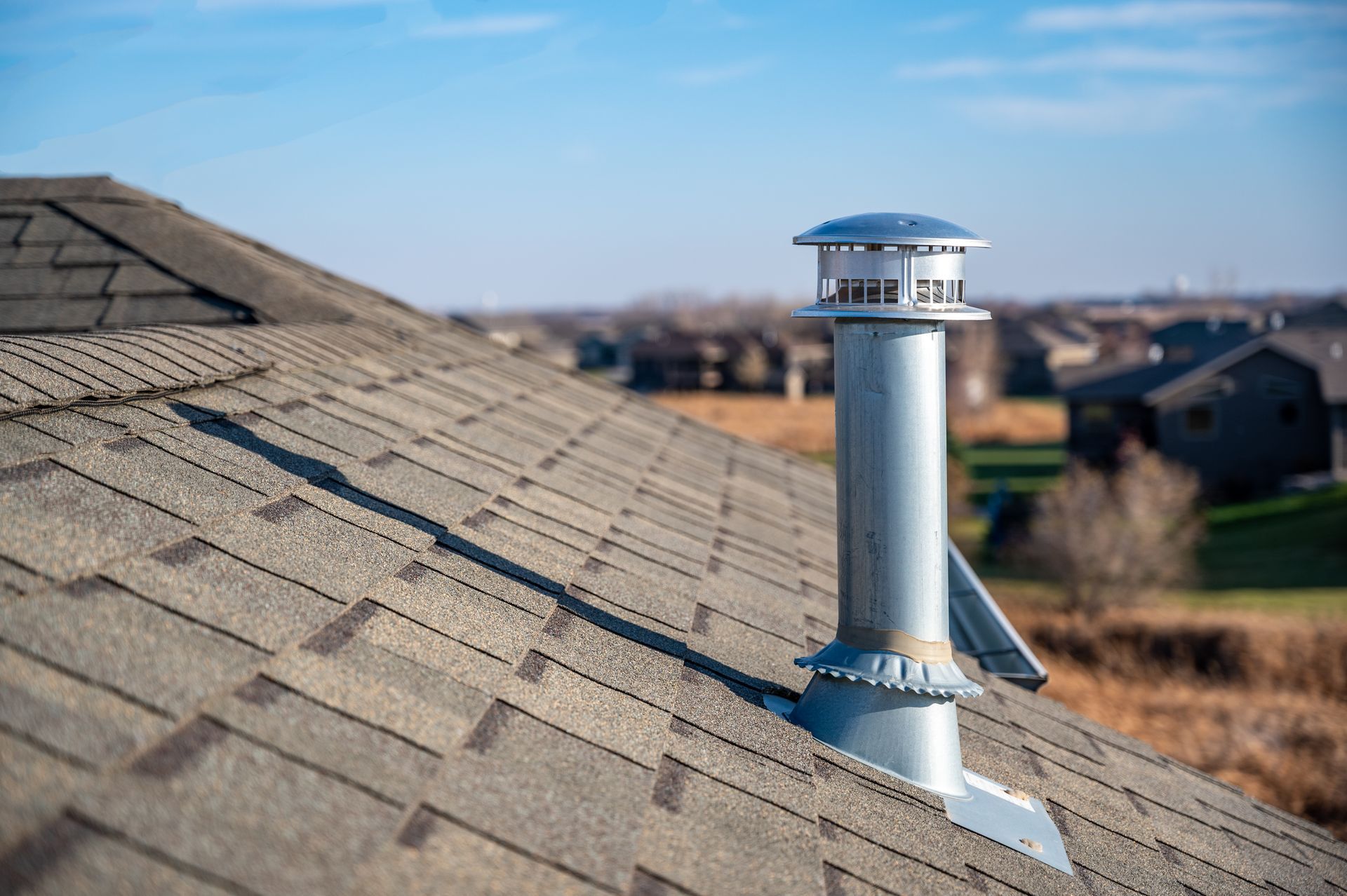 Conduit de ventilation métallique sur un toit en bardeaux sous un ciel bleu, avec des maisons de banlieue en arrière-plan